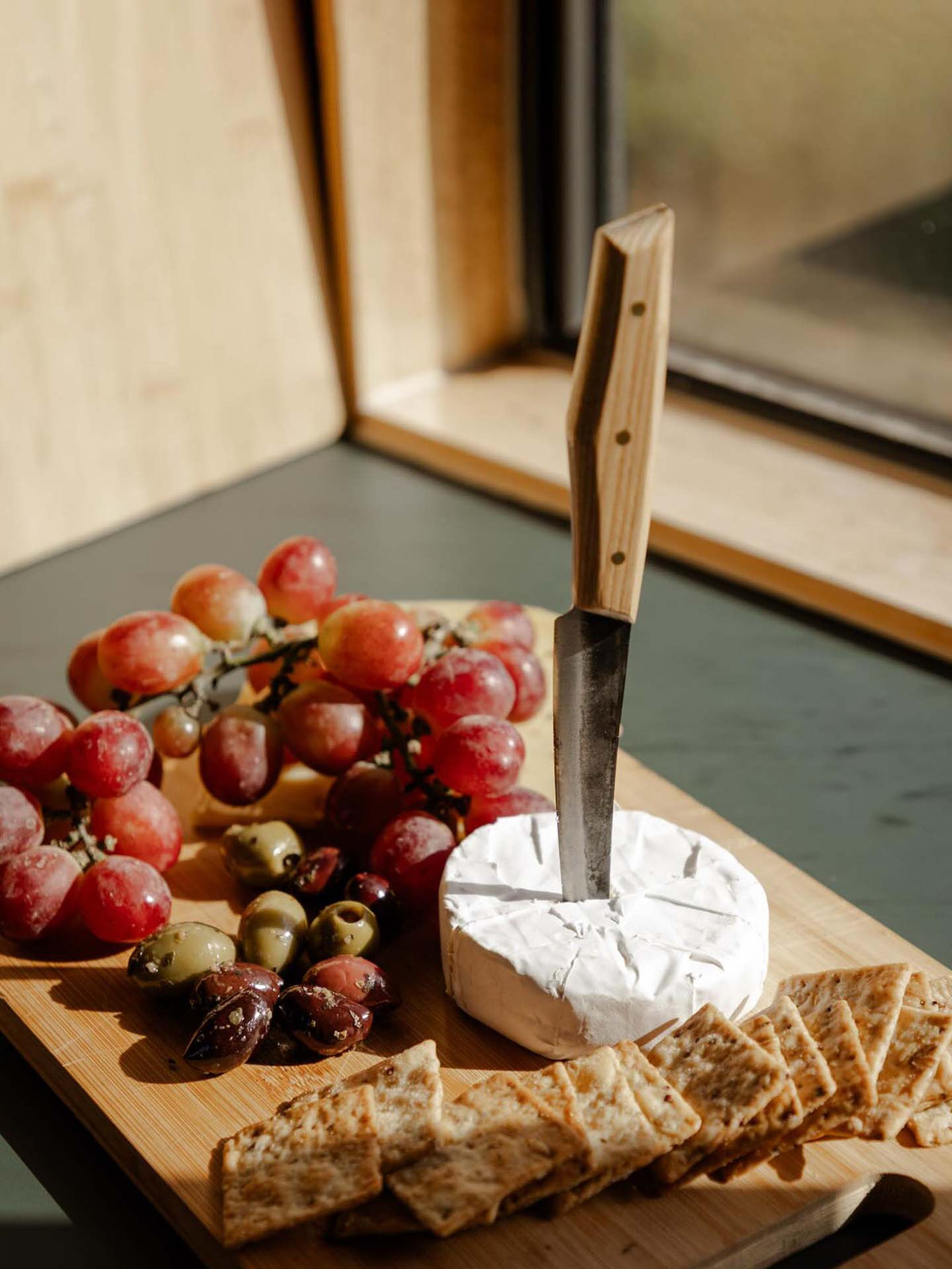 The №1 – Spread Knife is shown standing in a wheel of brie on a wooden board, surrounded by grapes, olives, and crackers near a window.