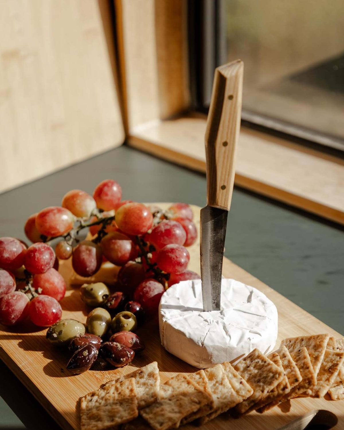 The №1 – Spread Knife is shown standing in a wheel of brie on a wooden board, surrounded by grapes, olives, and crackers near a window.