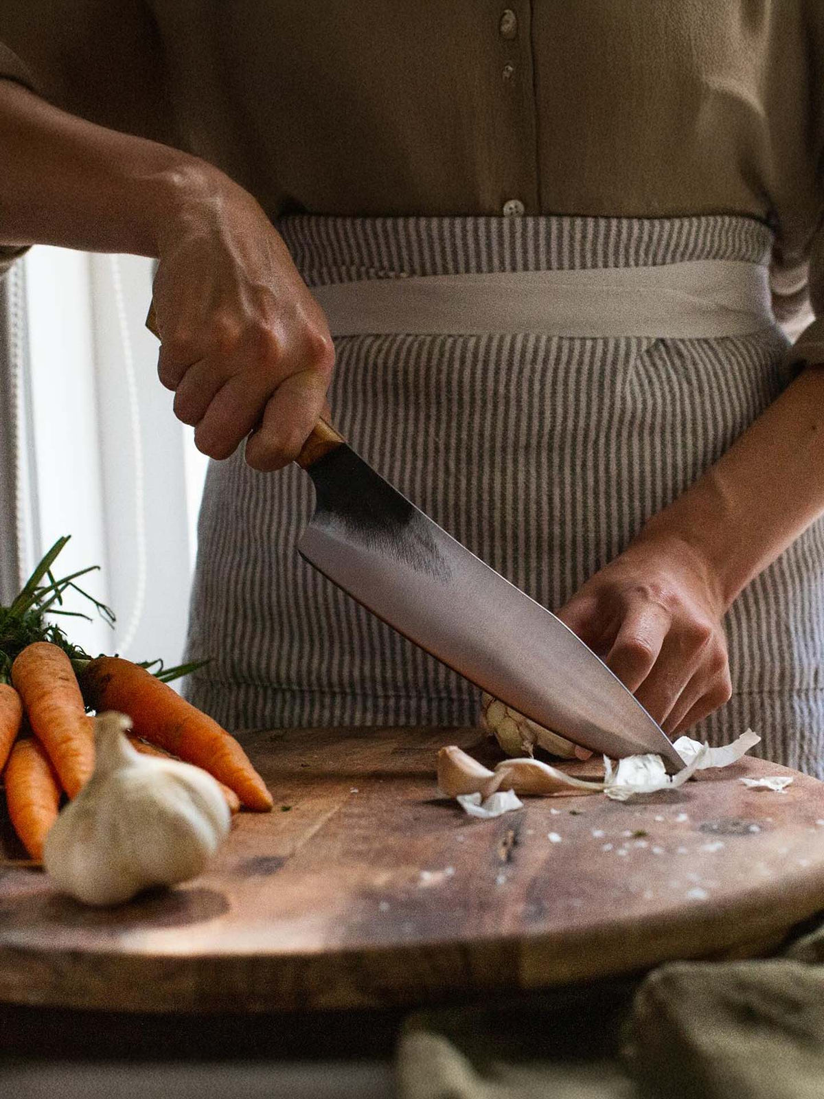 A person chops garlic with the №3 – Daily Knife on a wooden cutting board, wearing an apron, with carrots and a garlic bulb nearby.
