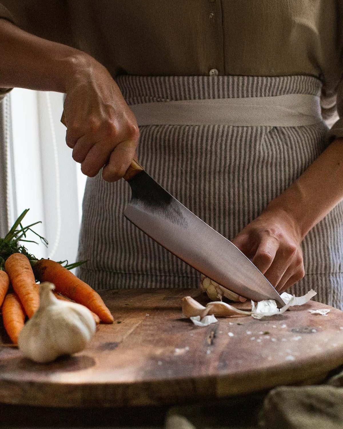 A person chops garlic with the №3 – Daily Knife on a wooden cutting board, wearing an apron, with carrots and a garlic bulb nearby.