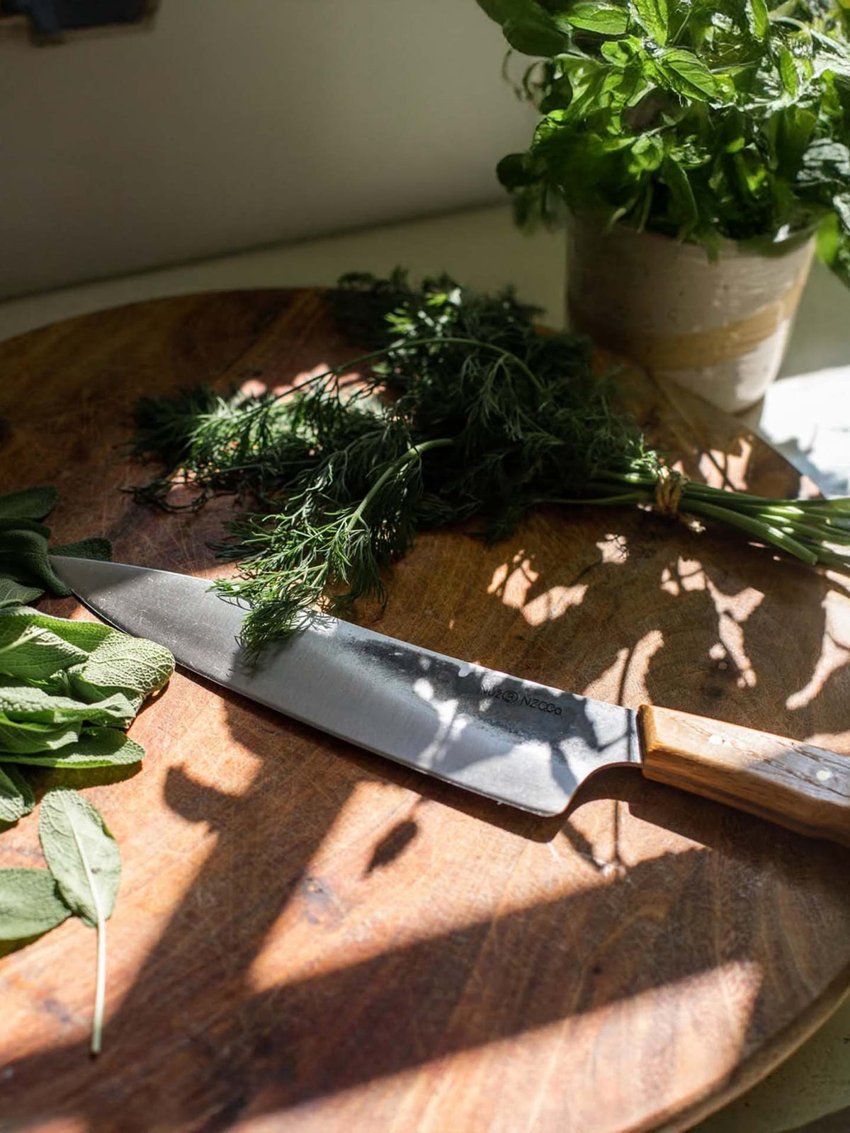 The №3 – Daily Knife, featuring a Swedish carbon steel blade, sits on a wooden cutting board with fresh sage and dill next to a sunlit potted herb plant.