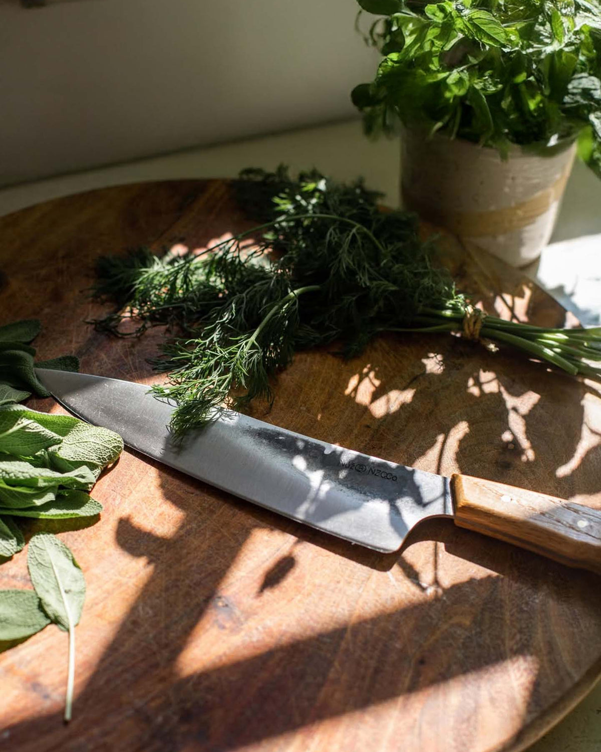 The №3 – Daily Knife, featuring a Swedish carbon steel blade, sits on a wooden cutting board with fresh sage and dill next to a sunlit potted herb plant.
