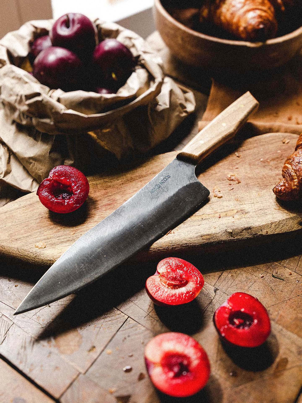 The №3 – Daily Knife rests on a wooden cutting board next to halved red plums, whole plums in a paper bag, and croissants atop a rustic table.