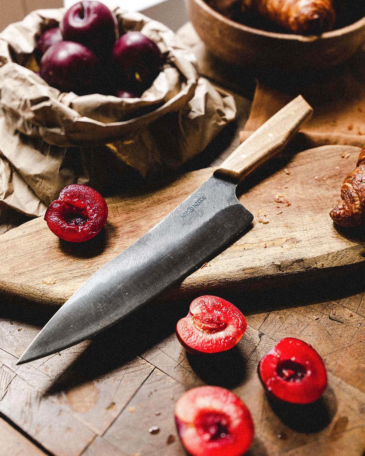 The №3 – Daily Knife rests on a wooden cutting board next to halved red plums, whole plums in a paper bag, and croissants atop a rustic table.