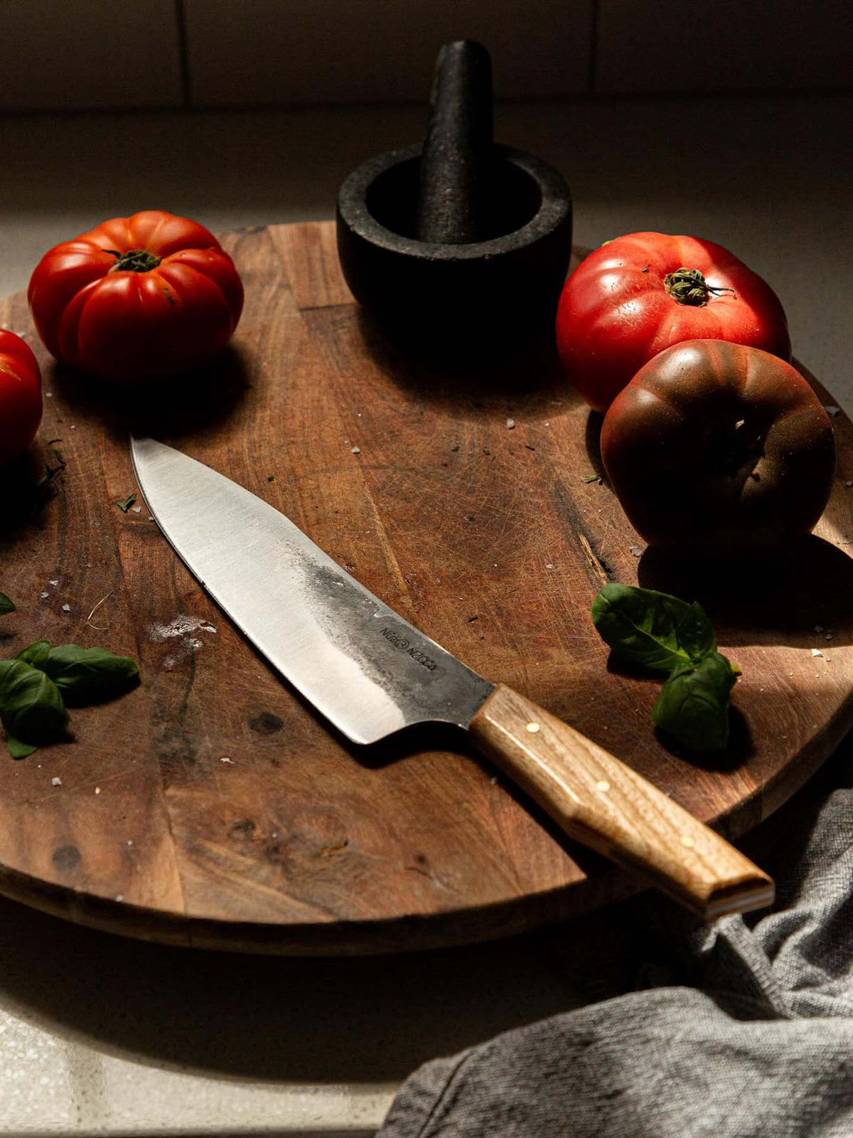 The №3 – Daily Knife rests on a wooden cutting board, surrounded by tomatoes, basil leaves, a mortar and pestle, and a gray kitchen towel.