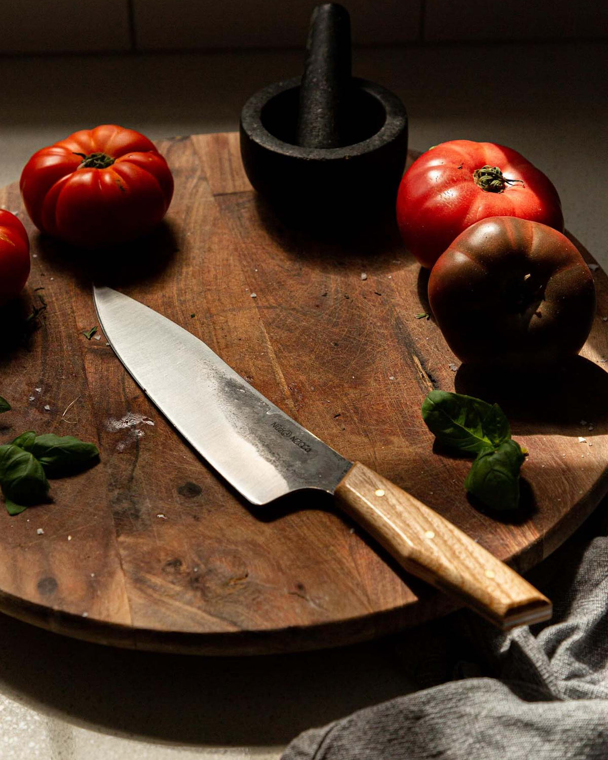 The №3 – Daily Knife rests on a wooden cutting board, surrounded by tomatoes, basil leaves, a mortar and pestle, and a gray kitchen towel.