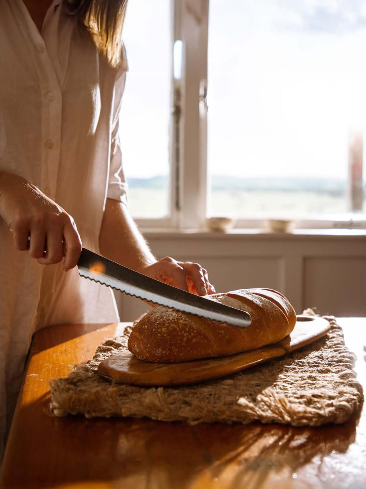 A person slices a loaf of bread on a wooden board with the №4 – Baker Knife, sunlight streaming through a window in the background.