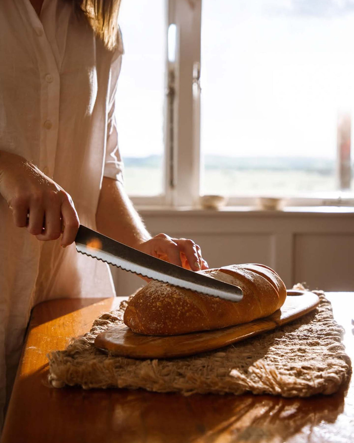 A person slices a loaf of bread on a wooden board with the №4 – Baker Knife, sunlight streaming through a window in the background.