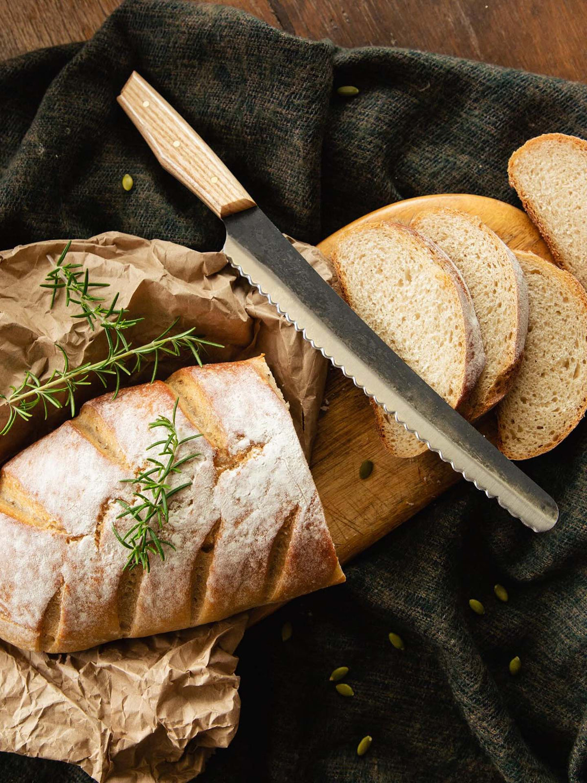 A loaf of crusty, handcrafted bread with rosemary on top and several slices rest on a wooden cutting board beside the №4 – Baker Knife.
