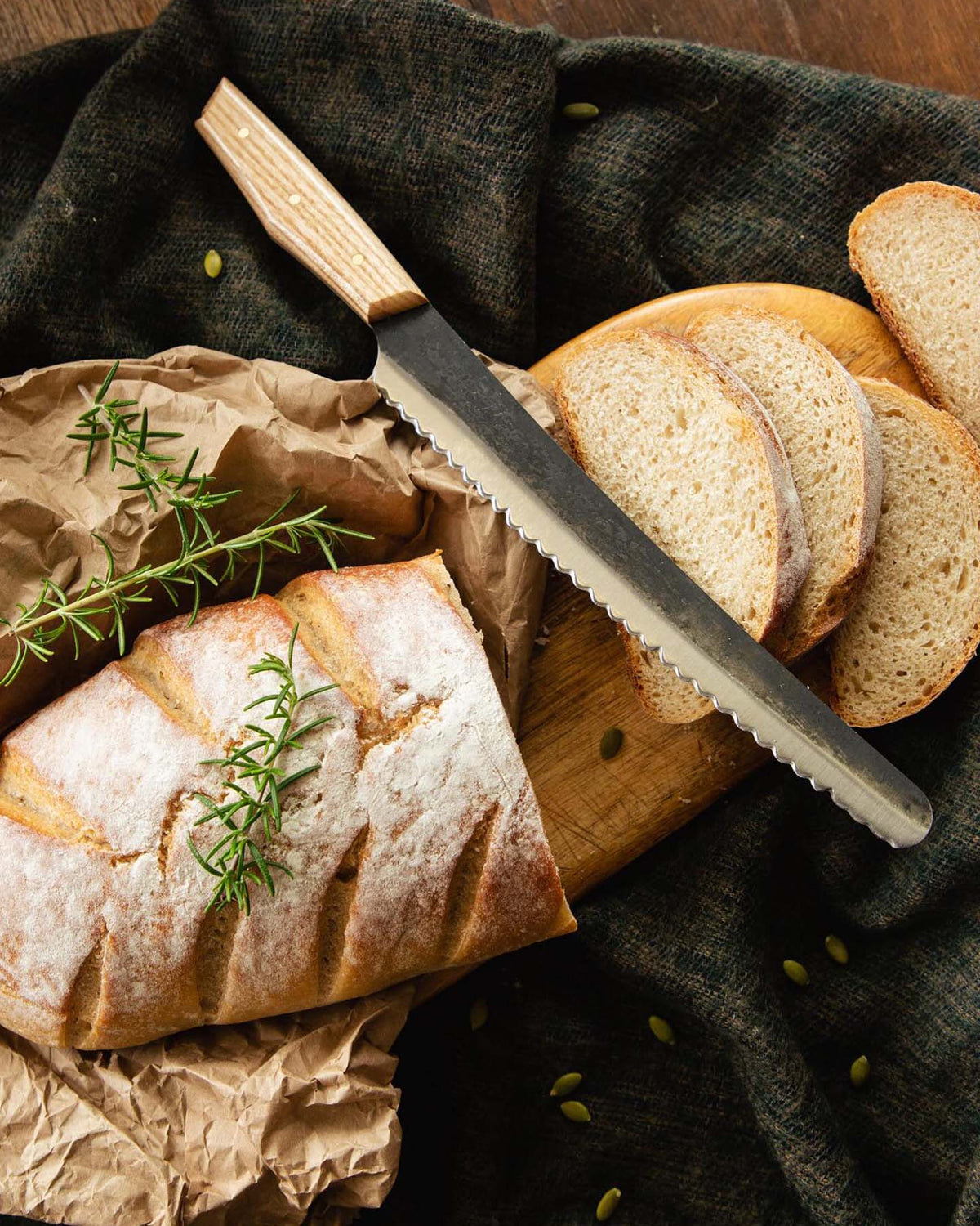 A loaf of crusty, handcrafted bread with rosemary on top and several slices rest on a wooden cutting board beside the №4 – Baker Knife.