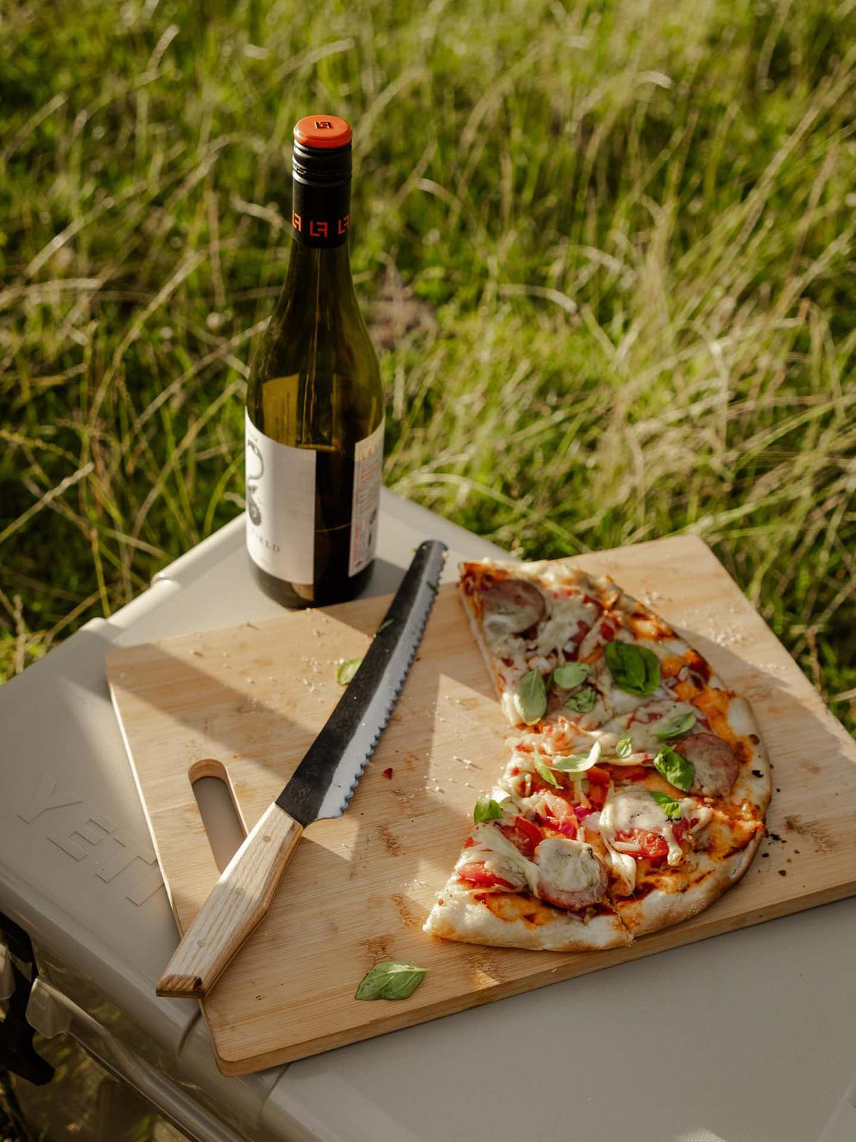 A partially sliced pizza with basil sits on a wooden board beside the №4 – Baker Knife and a wine bottle, arranged on a box outdoors in the grass.