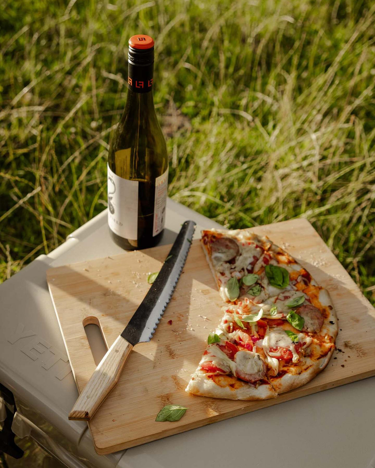 A partially sliced pizza with basil sits on a wooden board beside the №4 – Baker Knife and a wine bottle, arranged on a box outdoors in the grass.