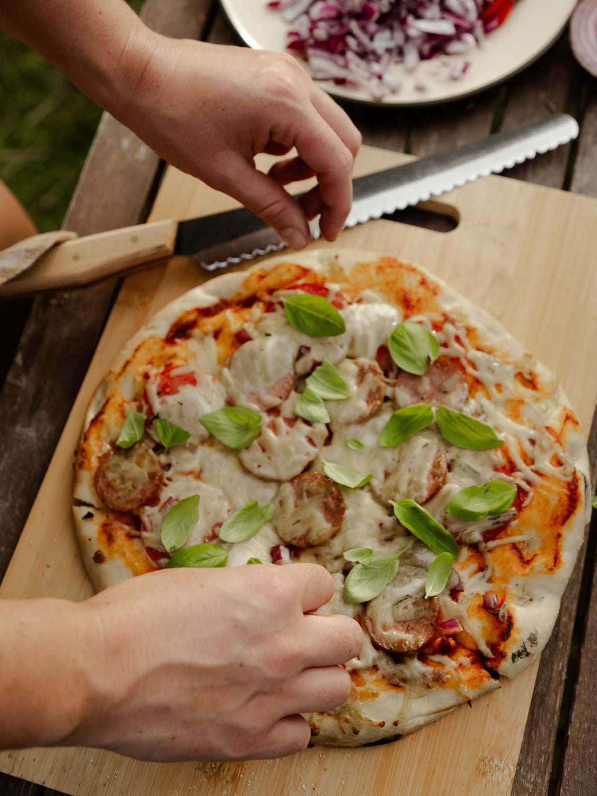 A person adds fresh basil to a homemade sausage and cheese pizza on a wooden cutting board, with the №4 – Baker Knife placed alongside.