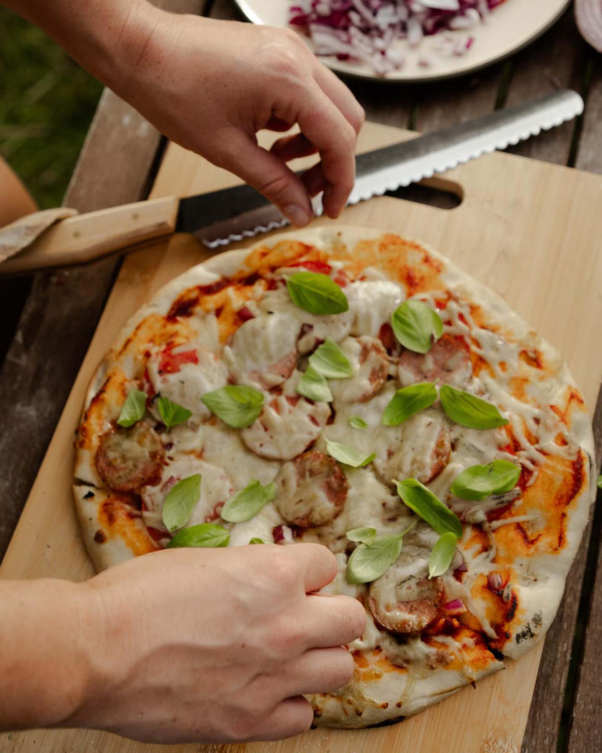 A person adds fresh basil to a homemade sausage and cheese pizza on a wooden cutting board, with the №4 – Baker Knife placed alongside.