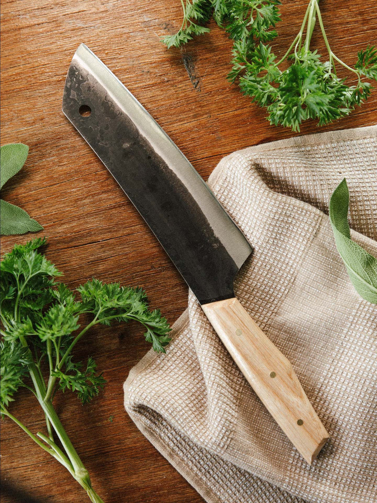 The №5 – Gourmet Knife, a large handcrafted Santoku with a wooden handle, rests on beige cloth amid fresh parsley and sage. Made from Swedish carbon steel, it combines functionality and elegance on any kitchen surface.