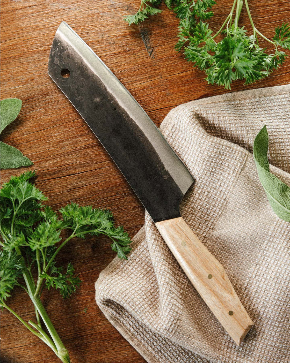 The №5 – Gourmet Knife, a large handcrafted Santoku with a wooden handle, rests on beige cloth amid fresh parsley and sage. Made from Swedish carbon steel, it combines functionality and elegance on any kitchen surface.