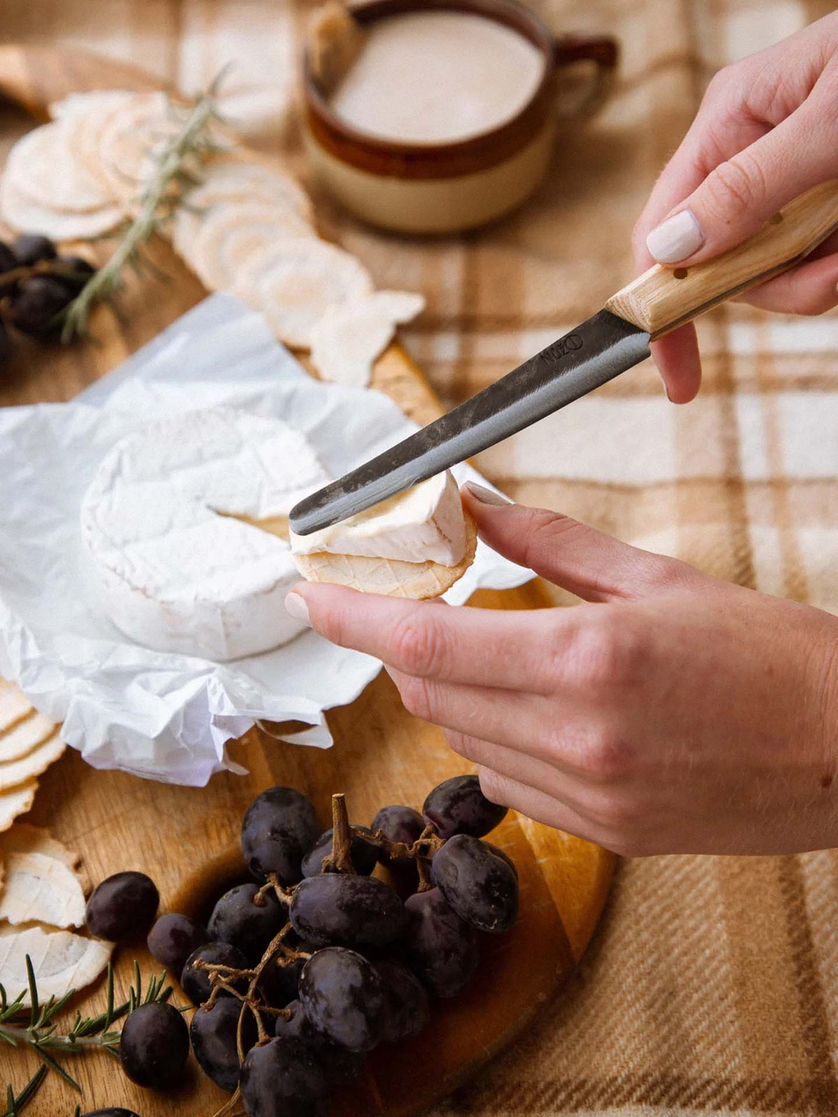 A person slices soft white cheese with the №1 – Spread Knife on a wooden board, surrounded by crackers, grapes, and fresh rosemary.