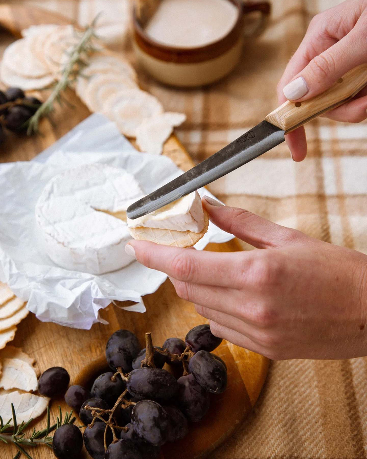 A person slices soft white cheese with the №1 – Spread Knife on a wooden board, surrounded by crackers, grapes, and fresh rosemary.