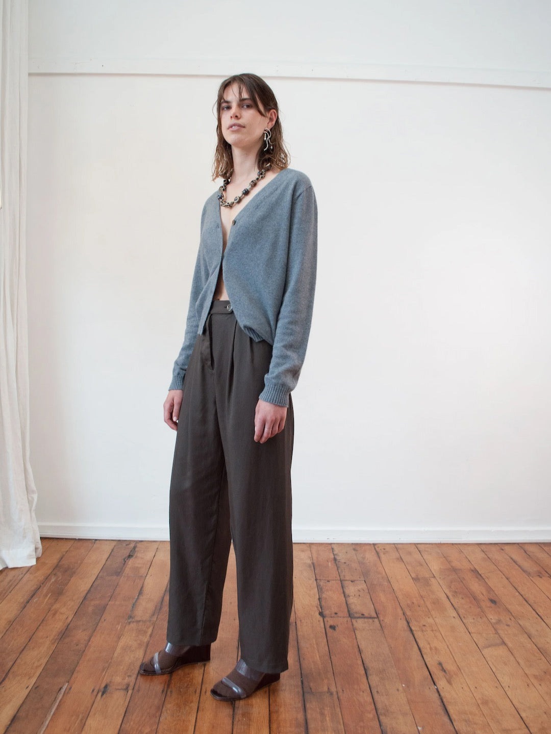 A woman is standing in a room wearing wide leg pants and an OVNA OVICH Kom Cardigan – Stone.