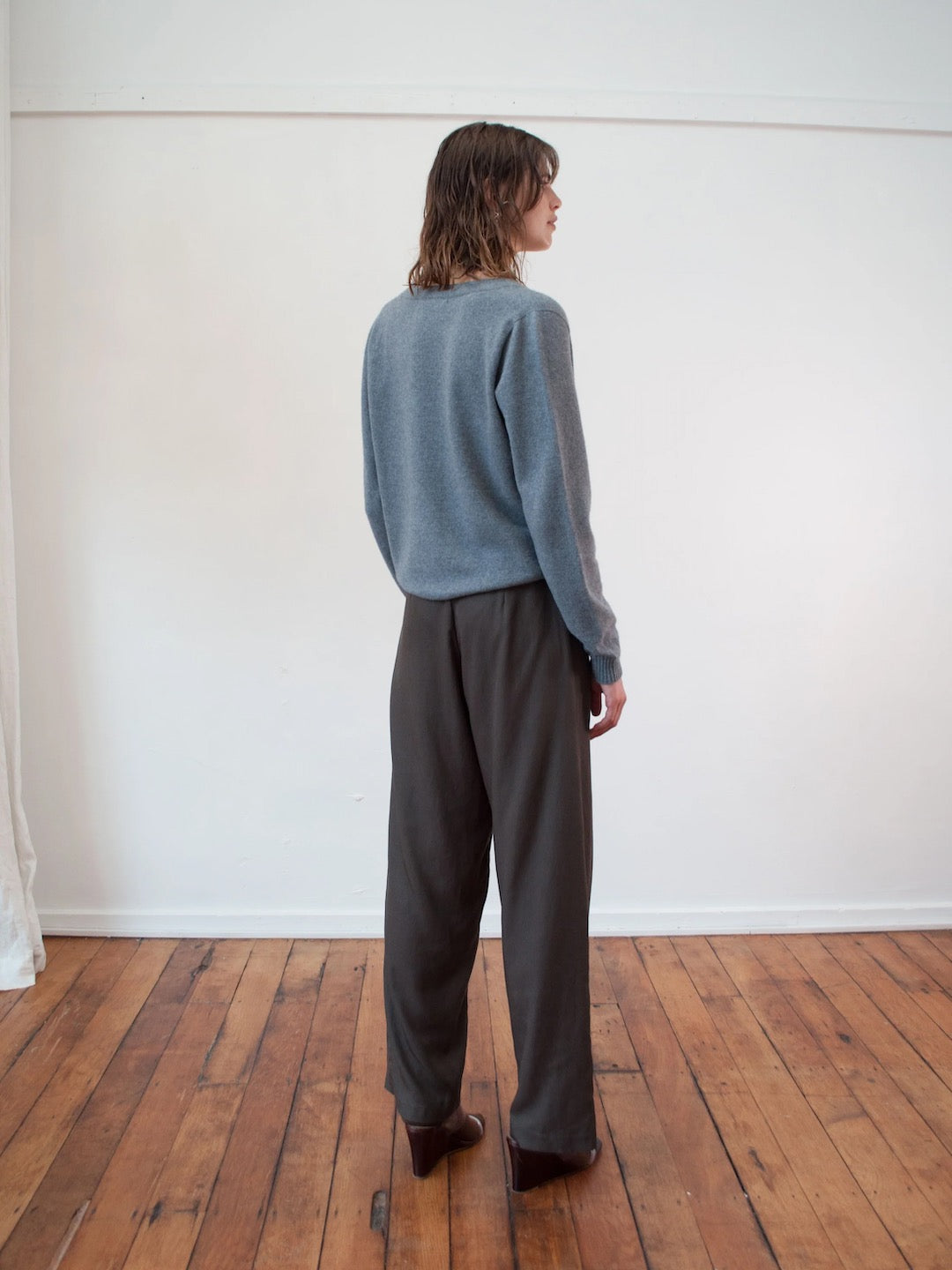 A woman is standing in a room wearing a Kom Cardigan - Stone by OVNA OVICH and trousers.
