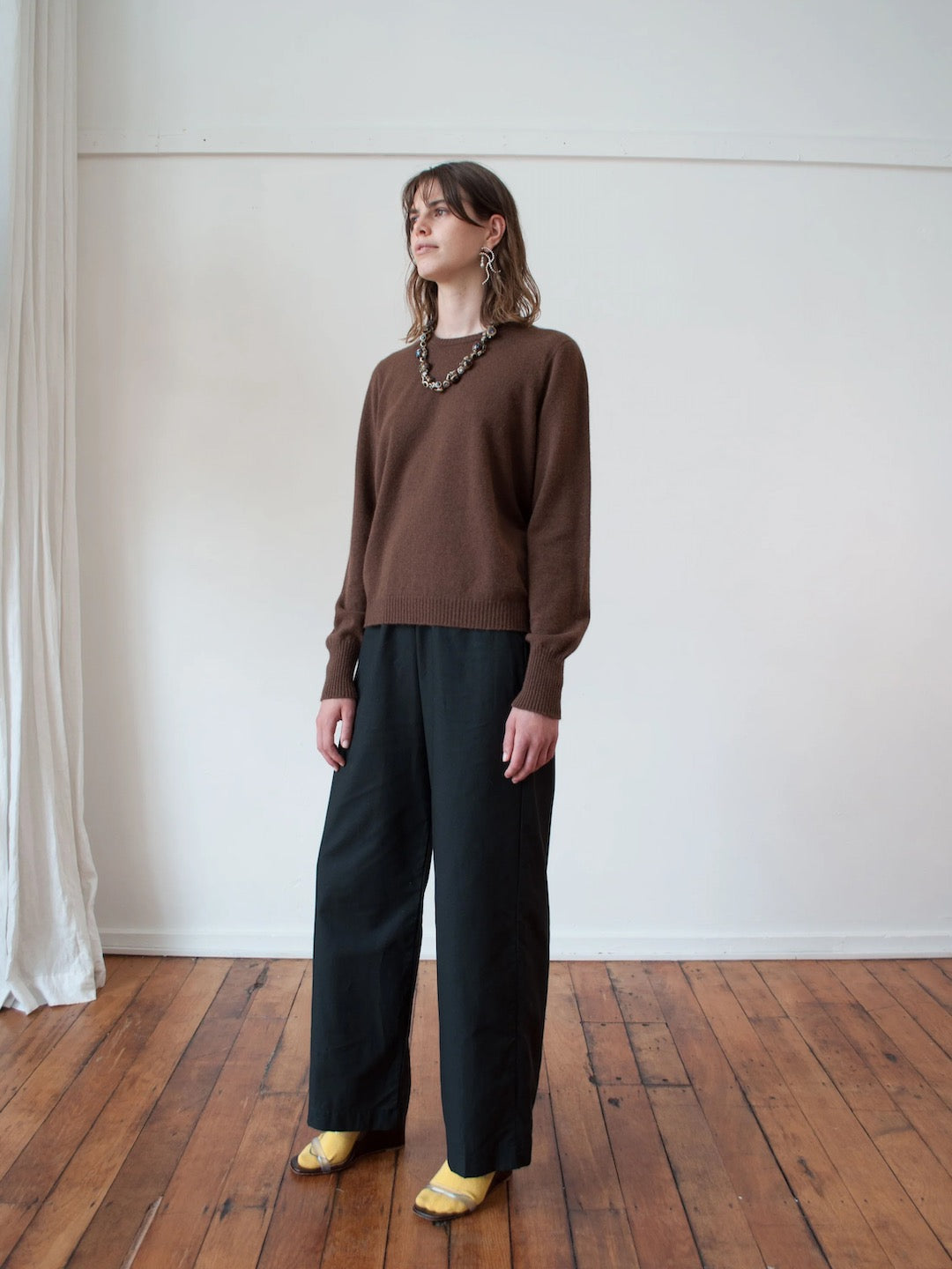 A woman standing in a room wearing an OVNA OVICH Kom Jumper – Mud and wide leg pants.