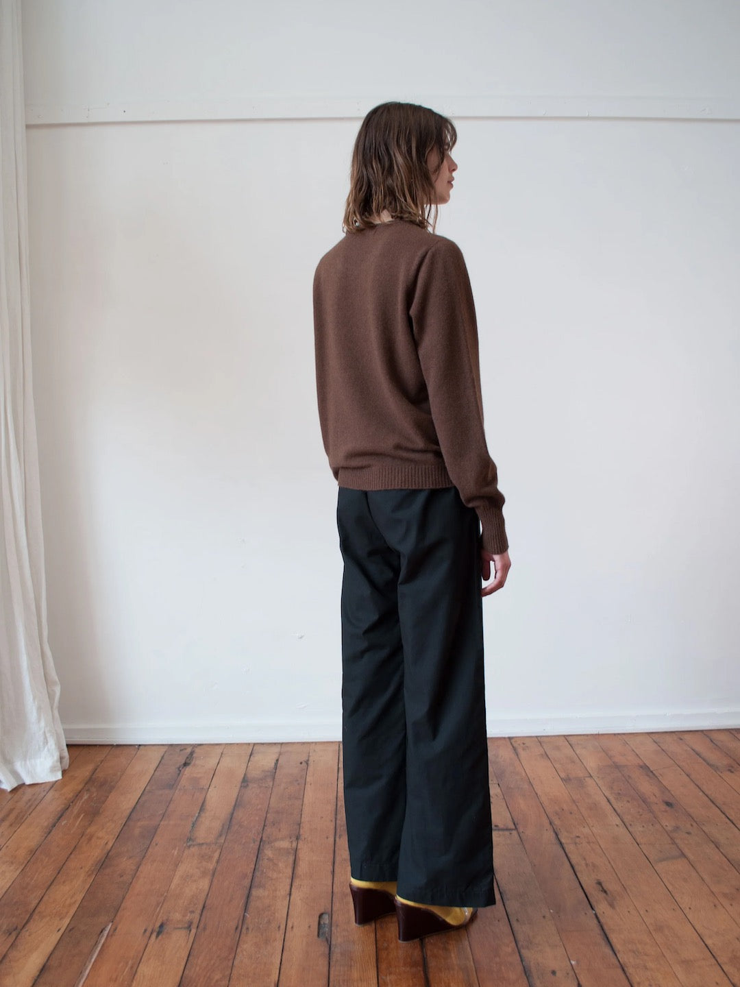 A woman is standing in a room wearing an OVNA OVICH Kom Jumper – Mud and wide leg pants.