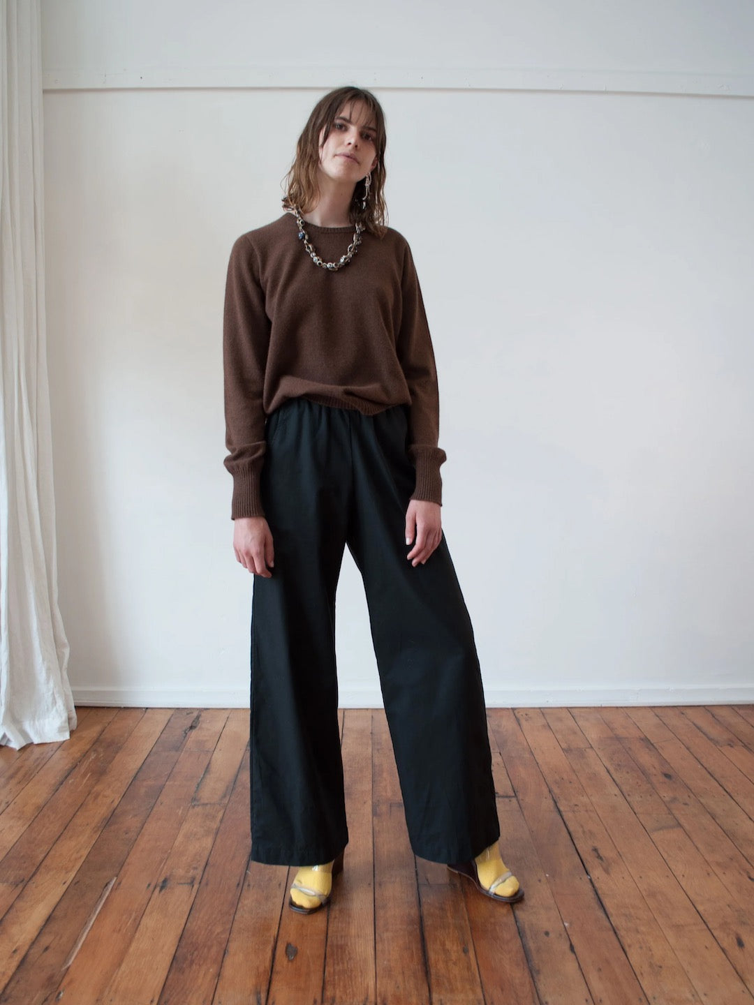 A woman is standing in a room wearing a Kom Jumper – Mud by OVNA OVICH and wide leg pants.