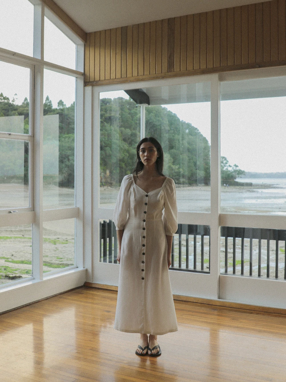 A sustainable Virginia Dress - Ice White designed by OVNA OVICH is worn by a woman standing elegantly in front of a window.