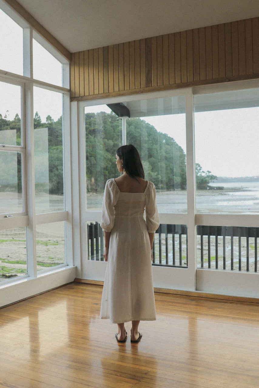 A sustainable woman in an OVNA OVICH Virginia Dress – Ice White standing in front of a large window.