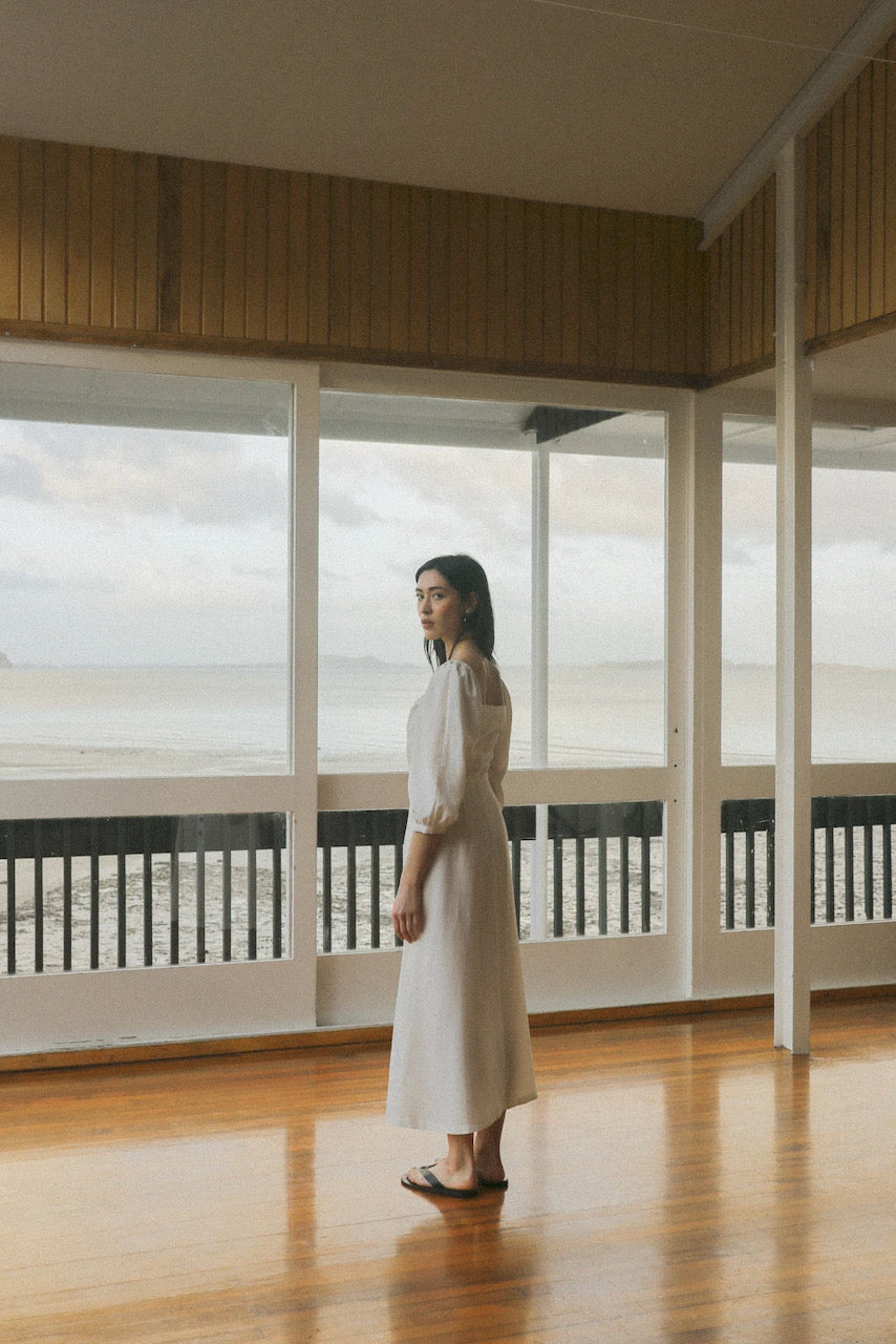 A sustainable OVNA OVICH-clad woman standing in a room with a view of the ocean, wearing the Virginia Dress - Ice White.