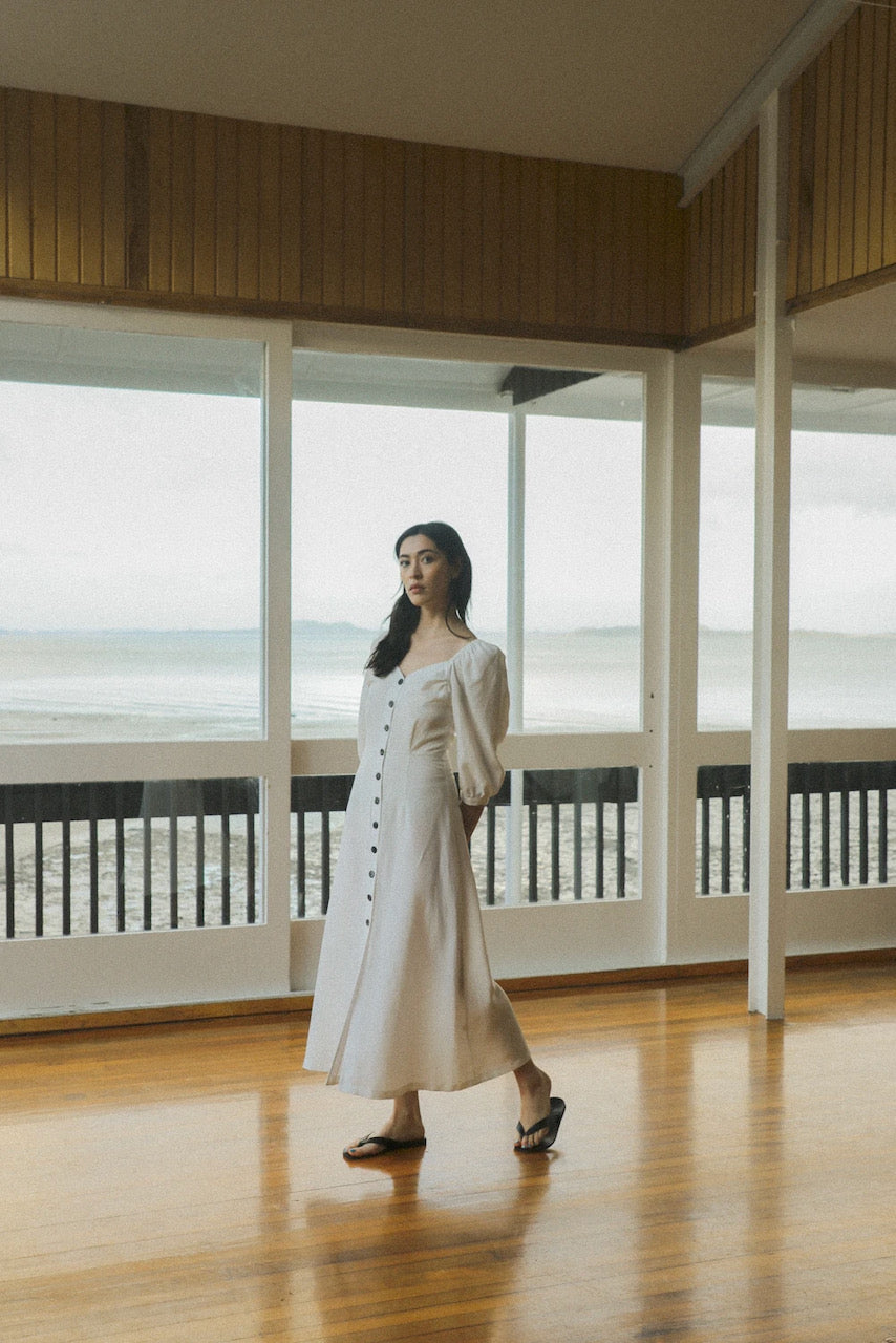 A sustainable woman wearing the Virginia Dress - Ice White by OVNA OVICH, standing gracefully on a wooden floor.