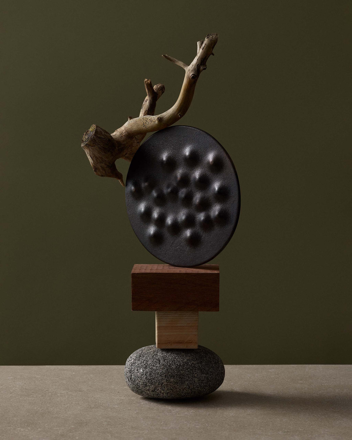A balanced sculpture consisting of a stone base, two wooden blocks, a dark circular disc with indentations, and a piece of driftwood.