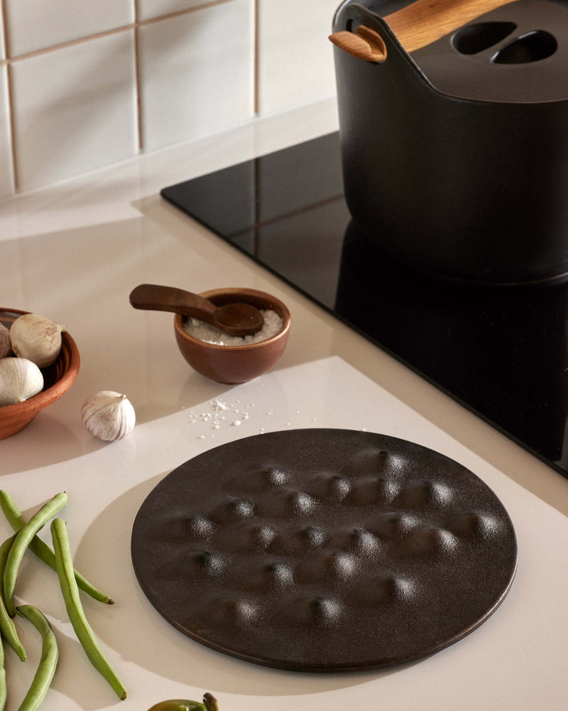 A round black cast-iron trivet with a textured, dimpled surface sits on a white kitchen counter, surrounded by cooking ingredients and a black stovetop pot.