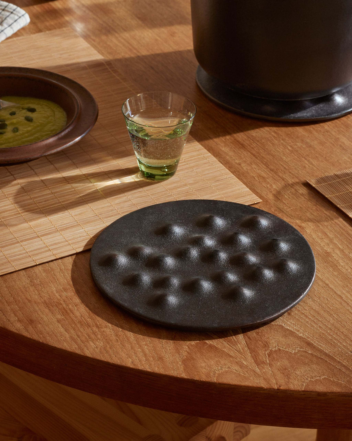 A round black textured trivet with raised dimples sits on a warm wooden dining table, styled with a small glass, bamboo placemats, and other tableware in natural sunlight.