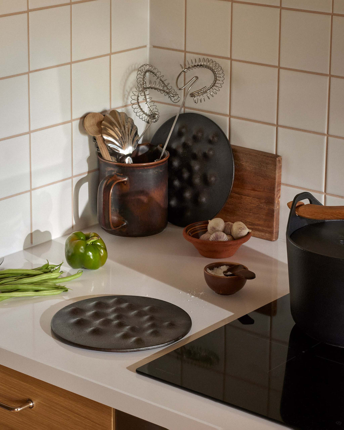 Kitchen counter with a cast-iron pan, ceramic mug holding whisks, green pepper, green beans, and a small bowl of garlic cloves.