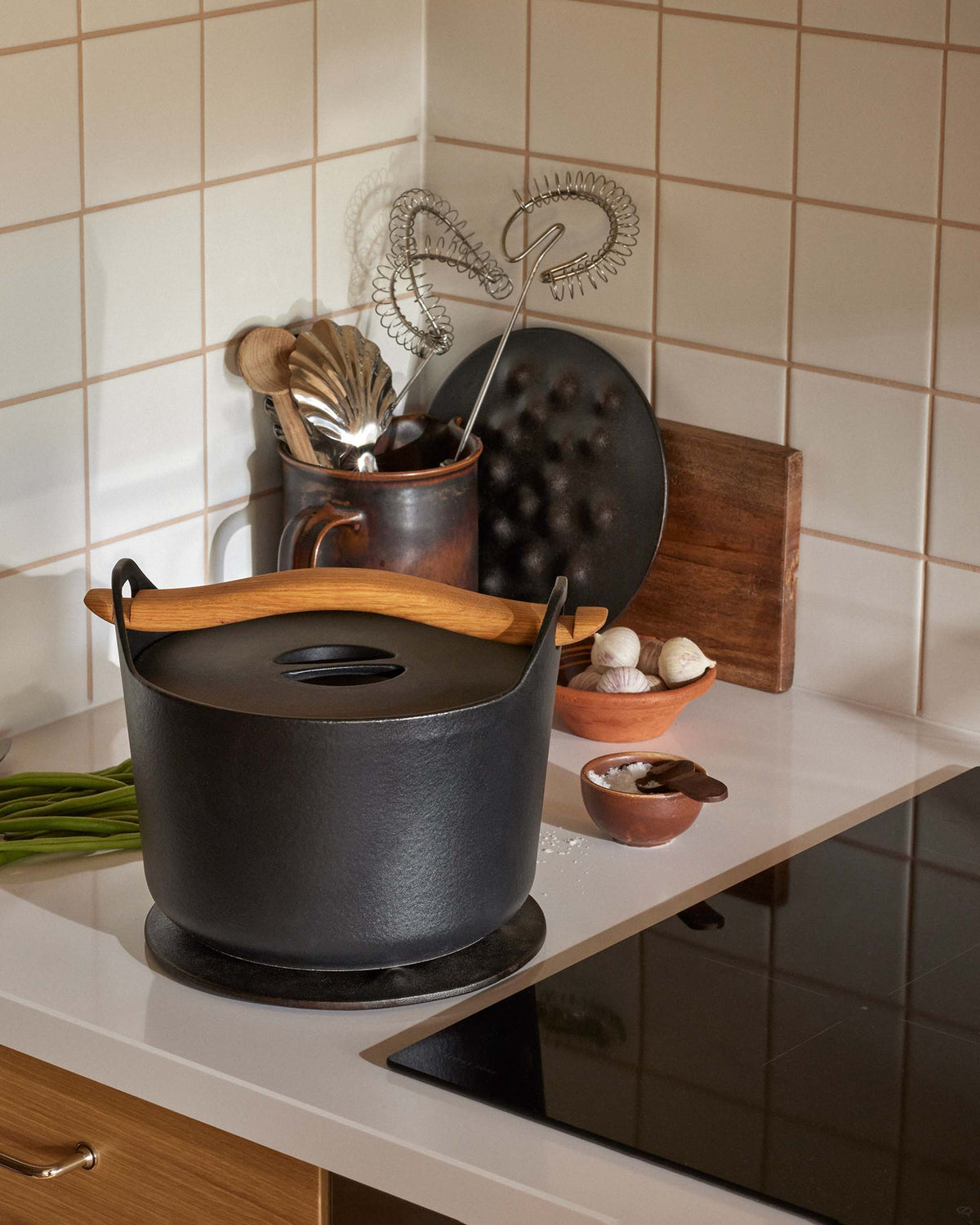A black cast iron pot with a wooden handle on a kitchen counter next to a stovetop, with kitchen tools and ingredients in the background.