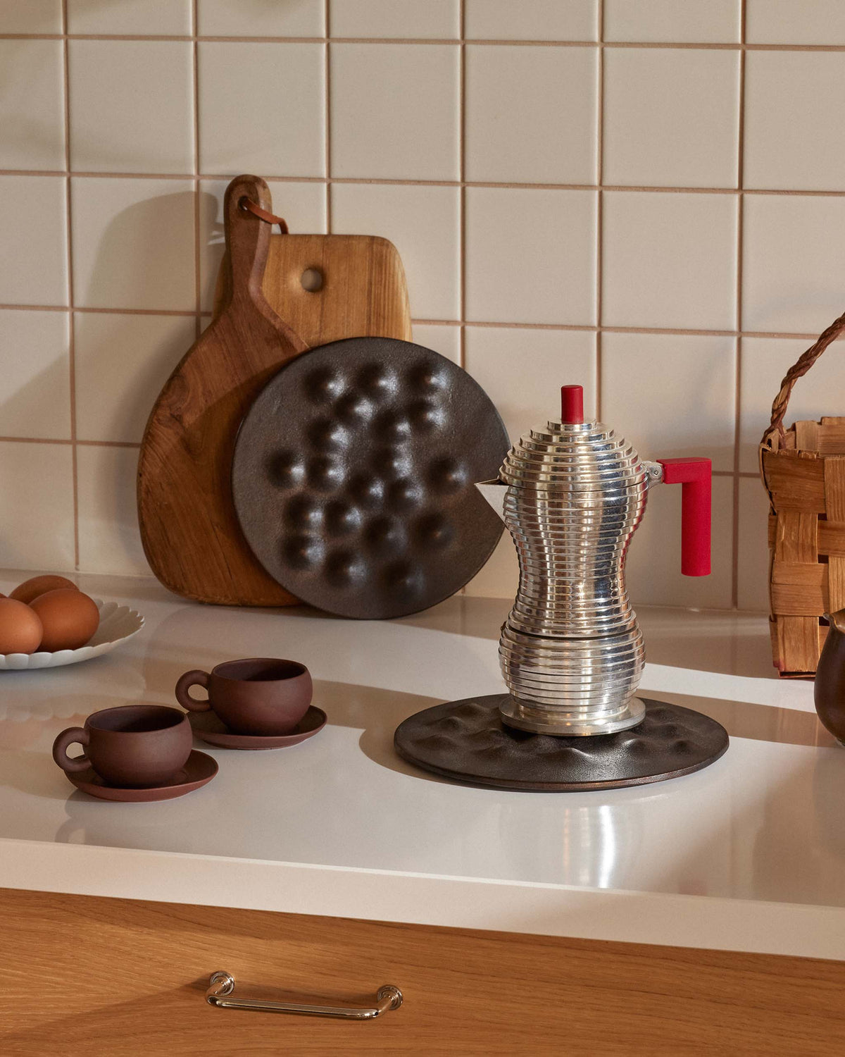 A ribbed silver moka pot with a red handle and knob, resting on a dark circular mat next to two brown cups and saucers.