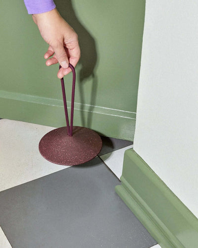 A hand is holding a maroon circular object with a loop handle, placed near the corner of a green-and-white checkered floor and green wall.