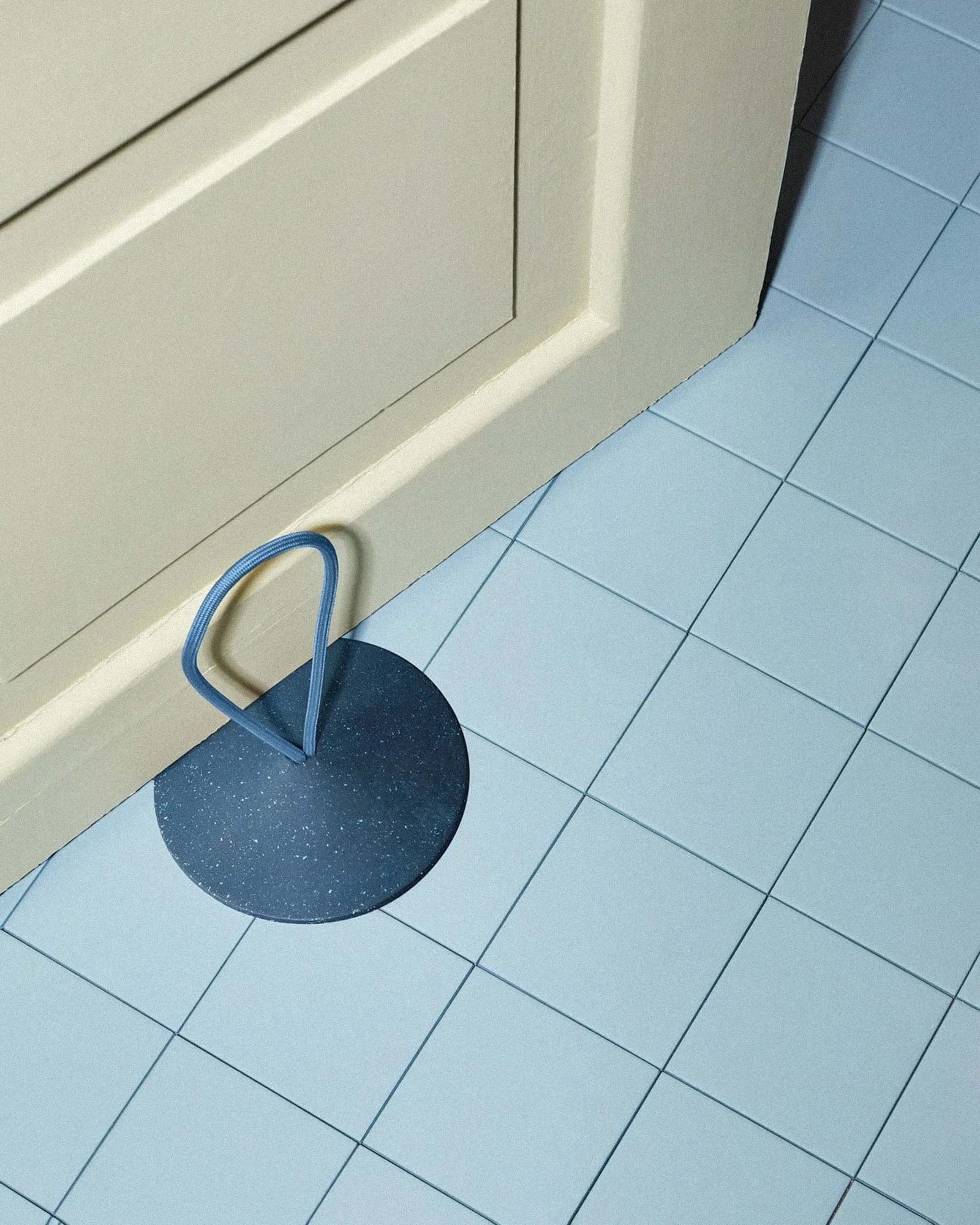 A round blue doorstop with a looped handle holds open a beige door on a light blue tiled floor.