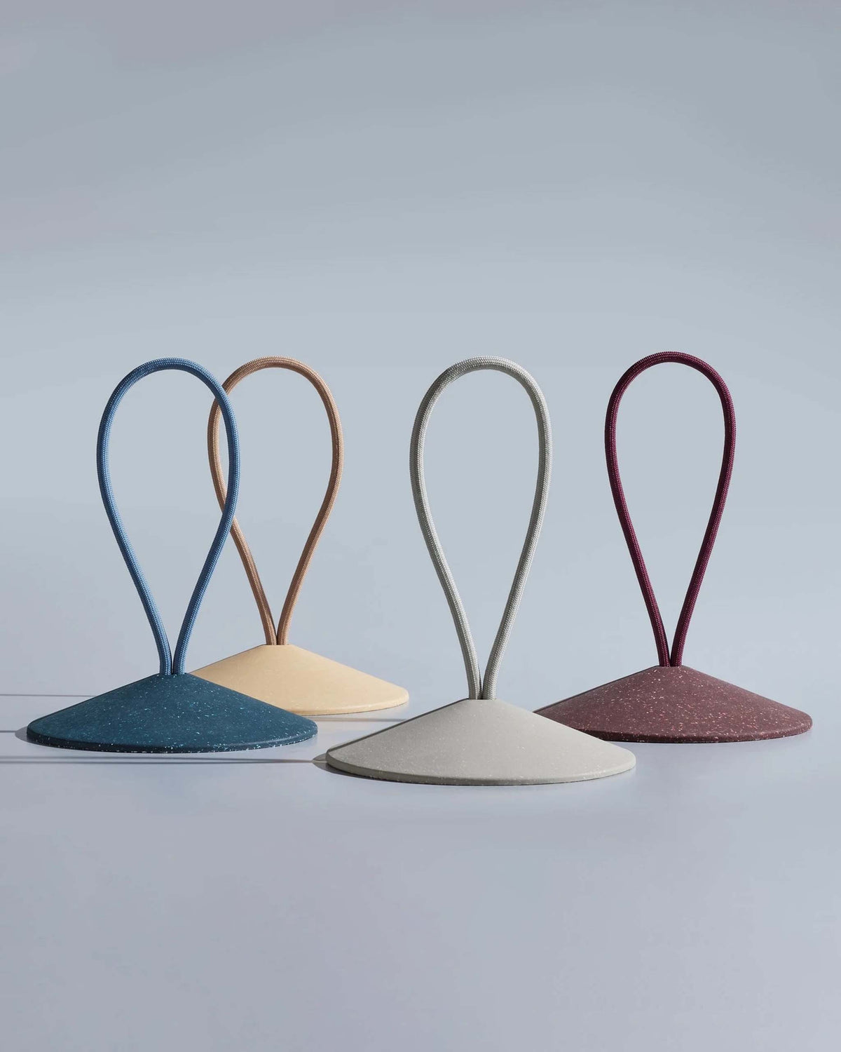 Four cone-shaped objects with large looped handles in blue, beige, gray, and maroon, are displayed upright against a plain, light gray background.