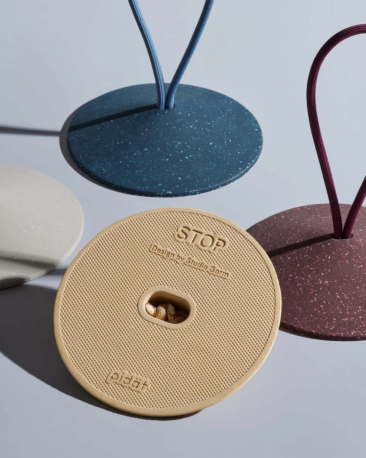 Four circular textured objects with a slot in one, labeled “STOP Design by Studio Born pidät,” and three standing bases with upright loops in blue, red, and beige on a light background.