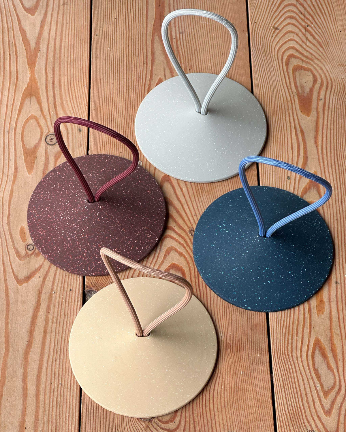Four round, speckled trivets with looped handles in beige, brown, gray, and blue arranged on a wooden surface.