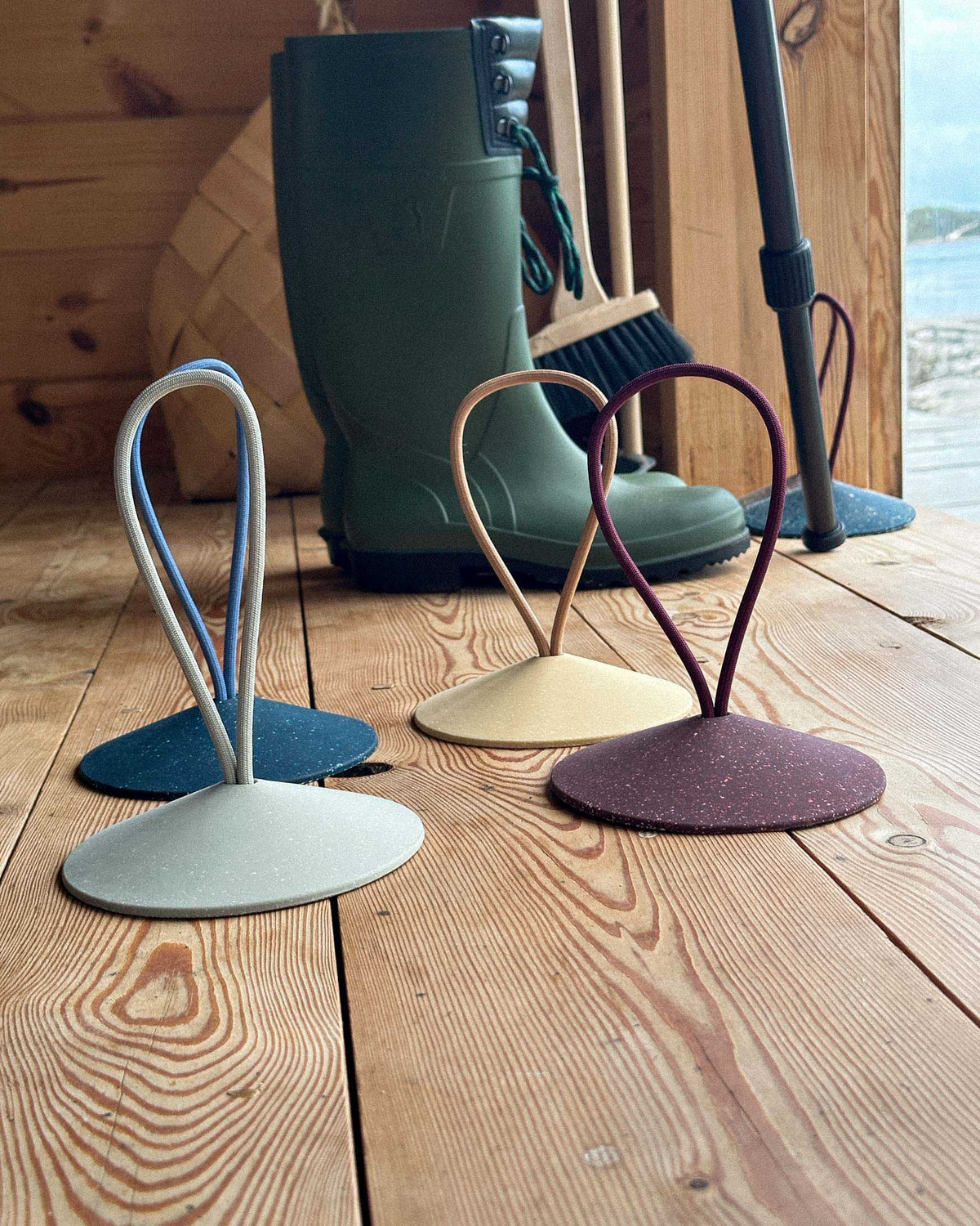 Three boot jacks in different colors are positioned on a wooden floor, with green rubber boots and cleaning tools in the background.