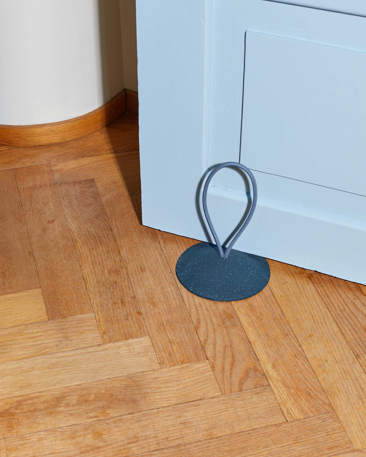 A blue metal doorstop with a looped handle holds a light blue door open on a wooden herringbone floor.