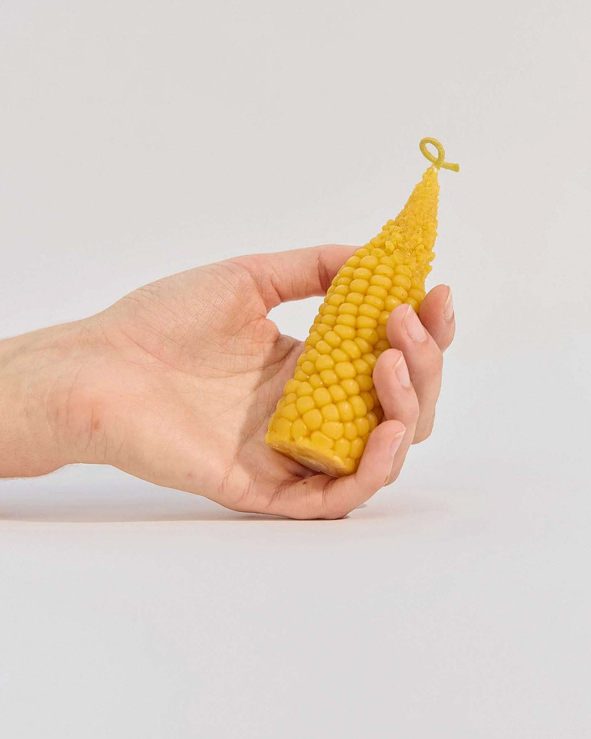 A hand holds the Sweetcorn Candle, a yellow ear-of-corn-shaped candle handmade from pure New Zealand beeswax, against a plain light background.