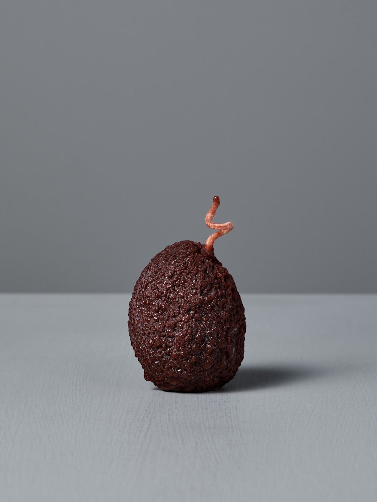 A dark green, textured Avocado Candle with a small, forked stem sits on a light gray surface against a plain gray background.