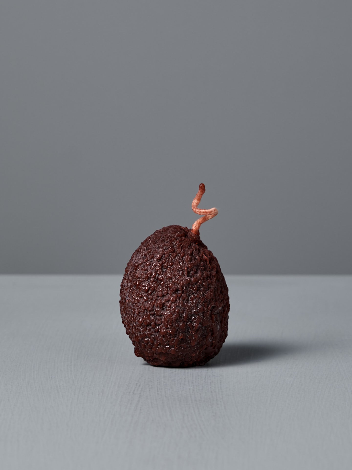 A dark green, textured Avocado Candle with a small, forked stem sits on a light gray surface against a plain gray background.