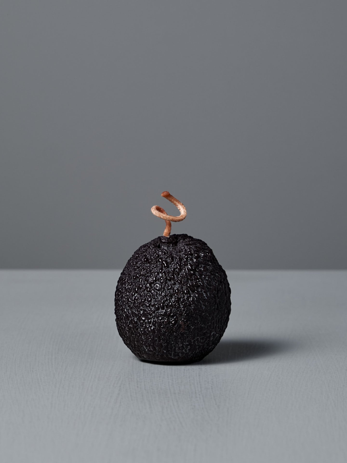 The Deep Purple Avocado Candle, handmade to resemble a ripe avocado with dark textured skin and a curly stem, stands upright on a gray surface against a plain gray background.