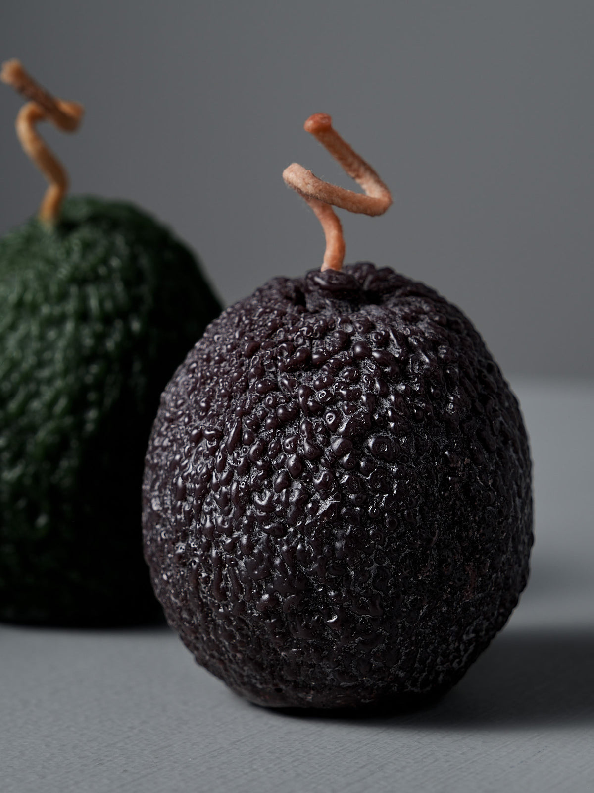 A Deep Purple Avocado Candle sits beside two avocados with twisted stems—one dark and ripe in front, one green and unripe behind—on a neutral surface.