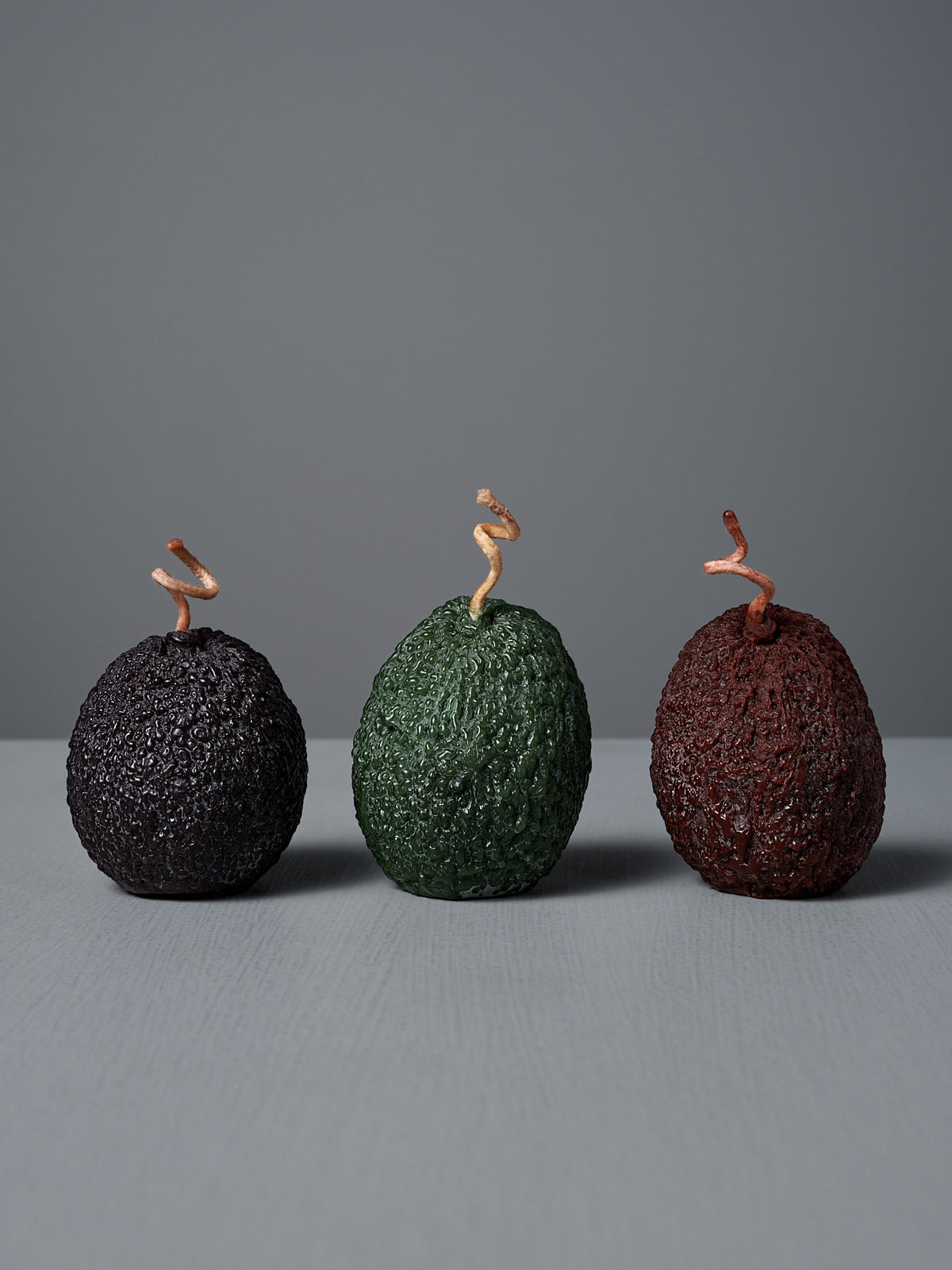 A Light Green Avocado Candle with a twisted stem is displayed on a gray surface against a gray background.